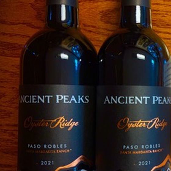 Two bottles of 2021 Ancient Peaks Oyster Ridge from Paso Robles, dark labels with copper script, standing on a wooden table