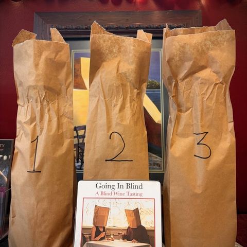 Three wine bottles hidden in numbered paper bags for a blind tasting, beside a Going In Blind event card